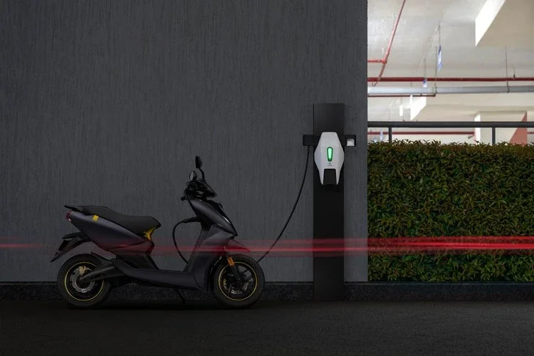 An electric scooter charging at a charging station in a parking garage with a gray wall and a green AI charging indicator.