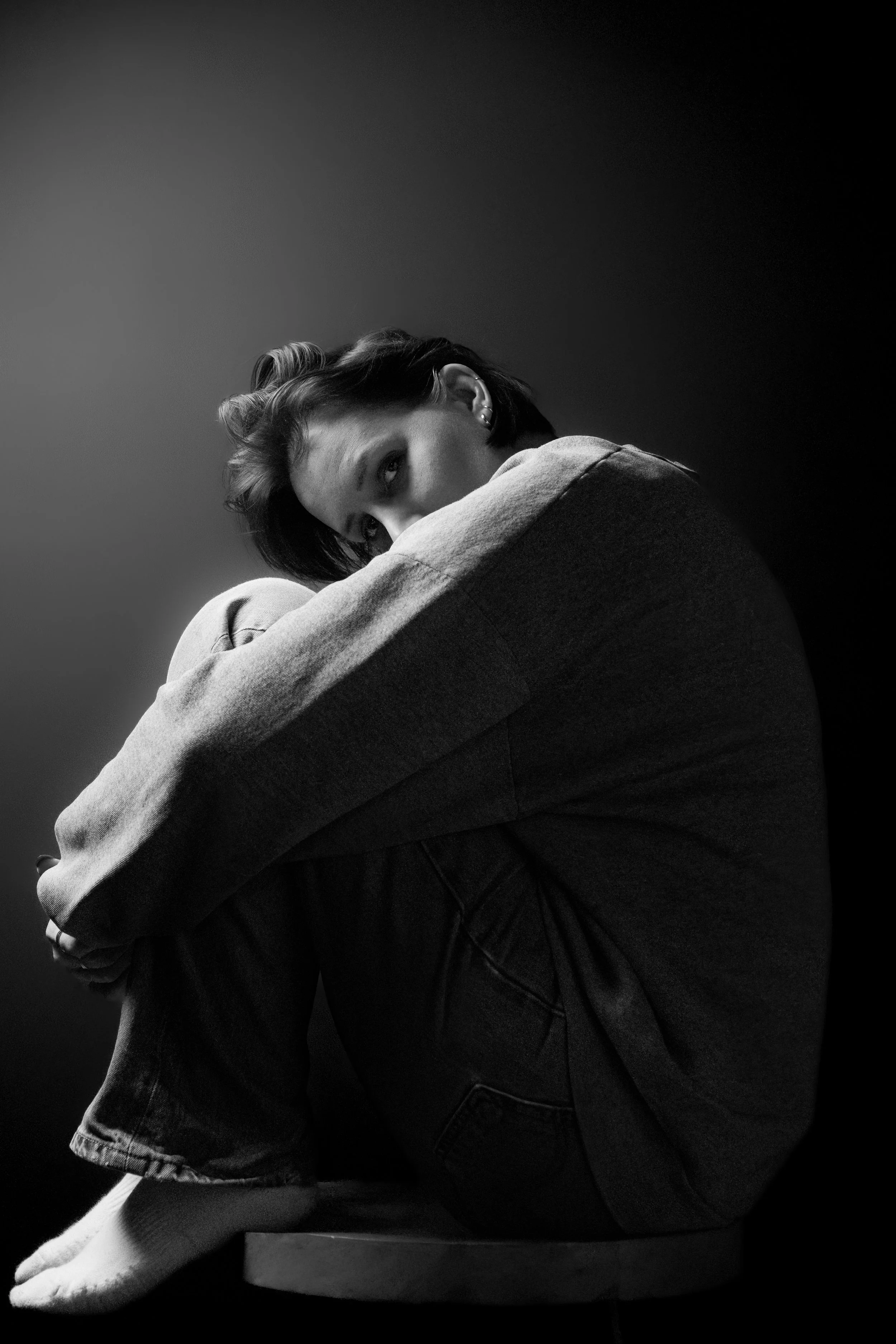 A woman with short hair sits on a stool, hugging her knees, looking at the camera in a black and white photograph by Susan Bradfield.  www.susanbradfield.co.uk