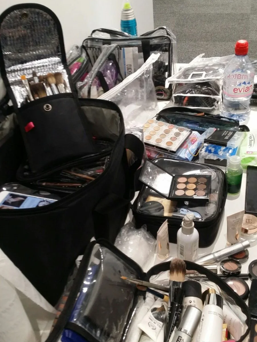 On set.  A table is laden with Professional makeup in bags.  Lots of different products, colours, brushes.