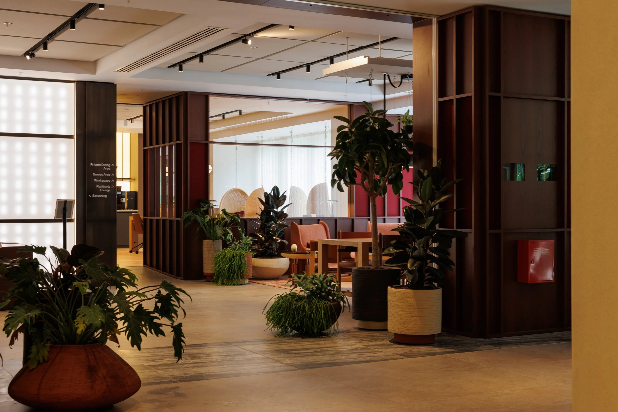 The image shows a chic hotel lobby with numerous green potted plants and cozy seating areas with chairs and tables. The ceiling has modern black track lighting, and there's a large white illuminated wall on the left side. The background features a hallway with glass chandeliers.