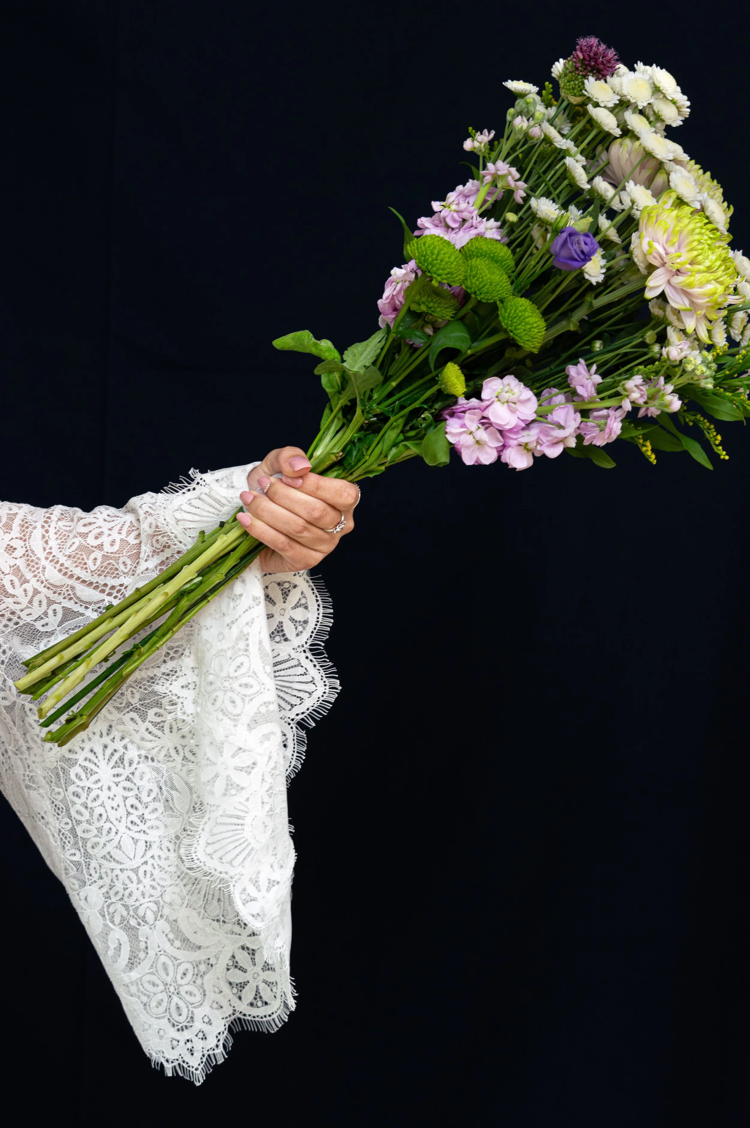 A hand holding a bouquet of assorted flowers against a black background. The arm wears a white lace sleeve. The bouquet includes pink, purple, yellow, and white flowers with green leaves.  Photography by Susan Bradfeld.  www.susanbradfield.co.uk