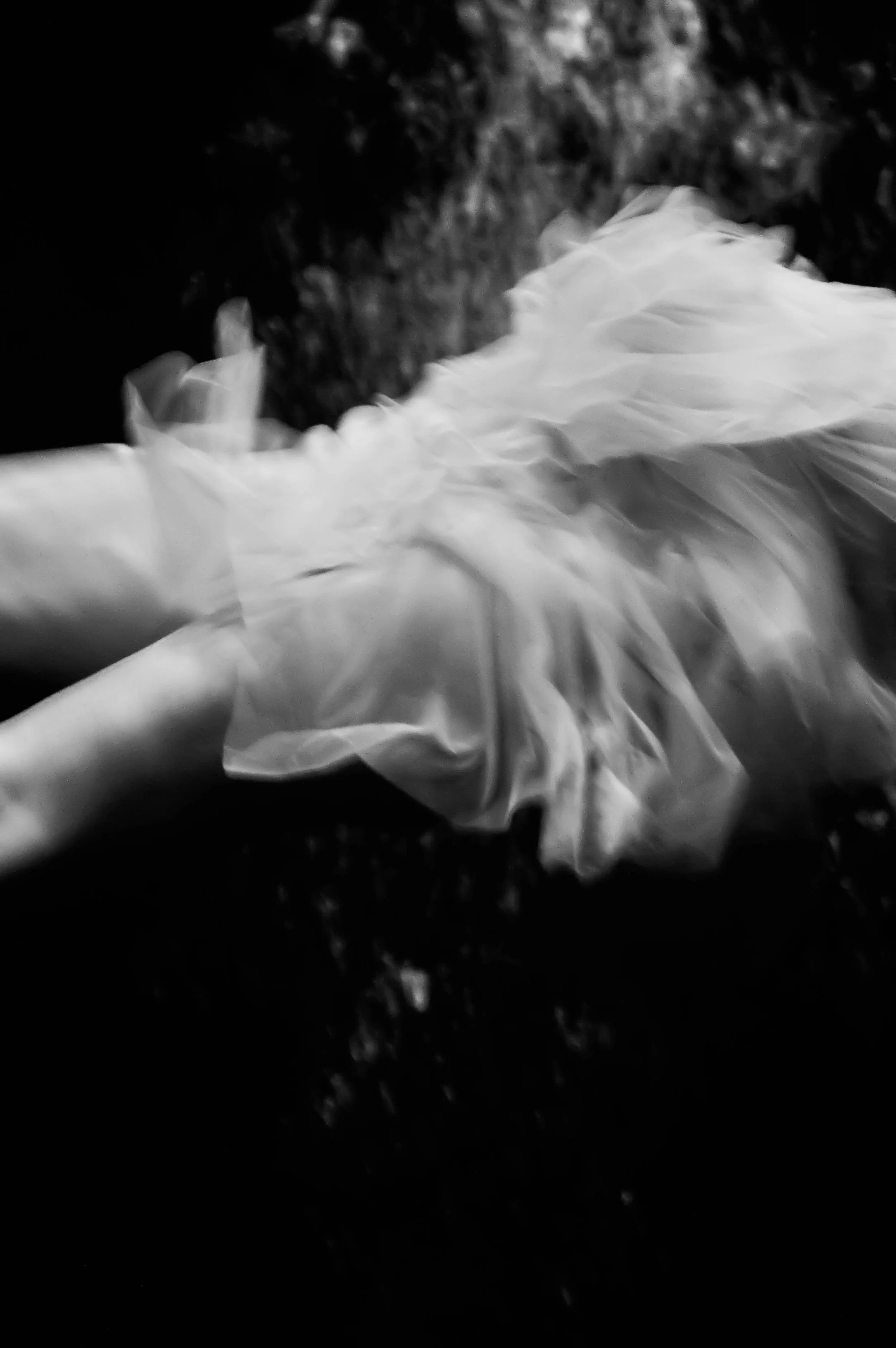 Black and white photograph taken by Susan Bradfield.  A girl floating underwater, her skirt swirls around as she sinks through the water, bubbles rising to the surface