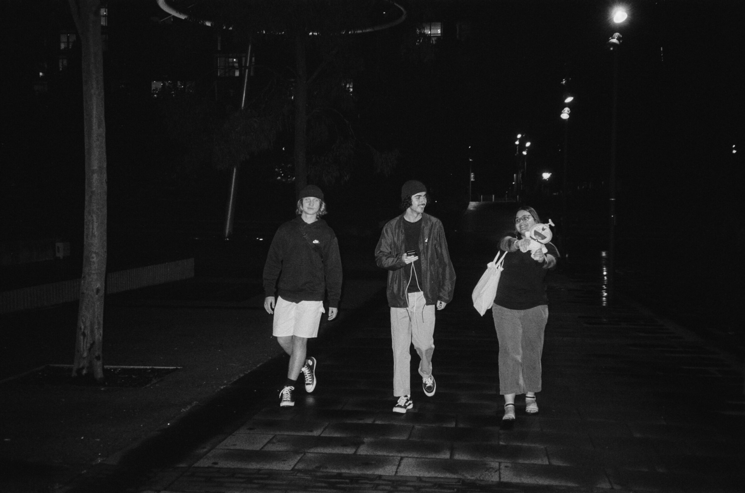 Three young people walking together at night on a lit walkway, one holding a plush toy and a tote bag, with trees and streetlights in the background.