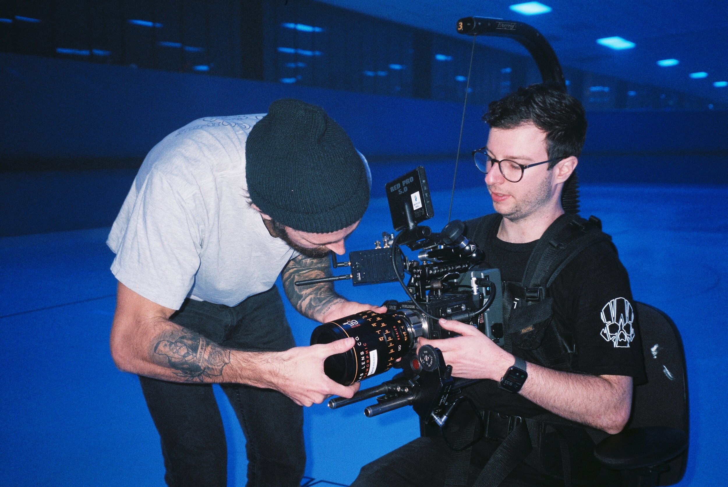 Two men are filming or inspecting a professional video camera indoors, with a background of blue lighting.