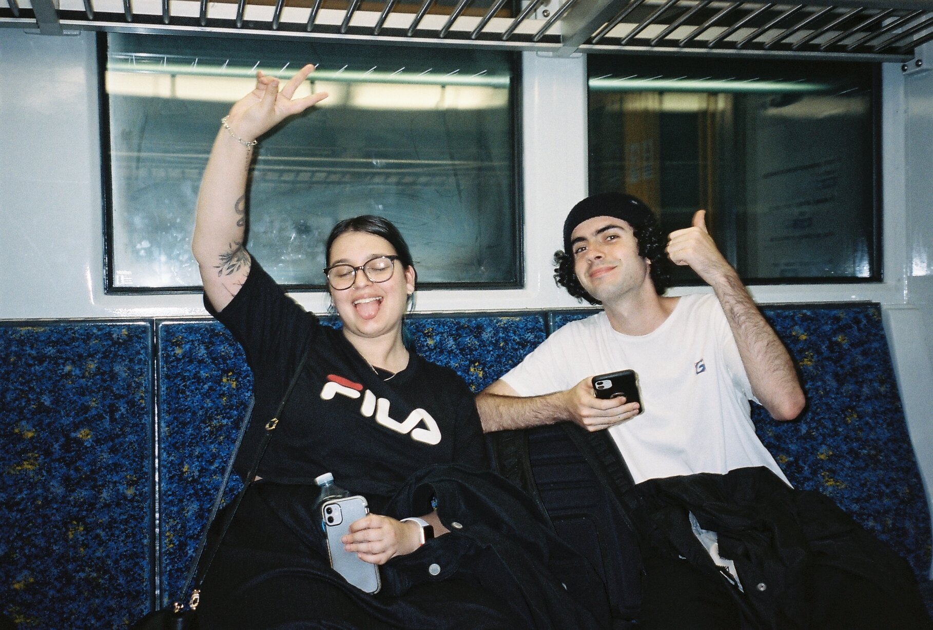 Two young people sitting on a bus bench, smiling and making gestures; the woman is wearing glasses and a FILA T-shirt, the man has curly hair and is wearing a white T-shirt, holding a smartphone.
