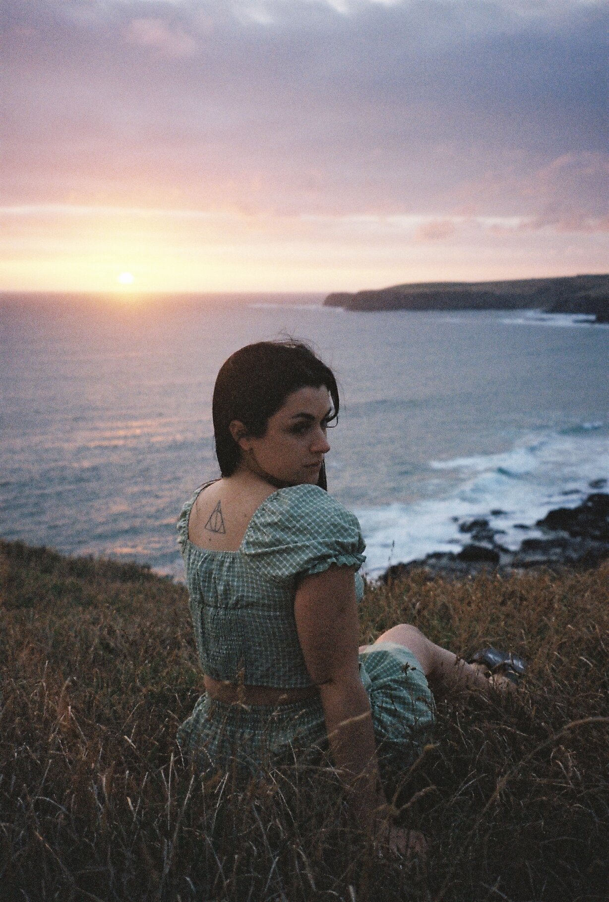 A woman with short dark hair sitting on tall grass at sunset near the ocean, with a tattoo of a sailboat on her shoulder, wearing a green checkered dress.