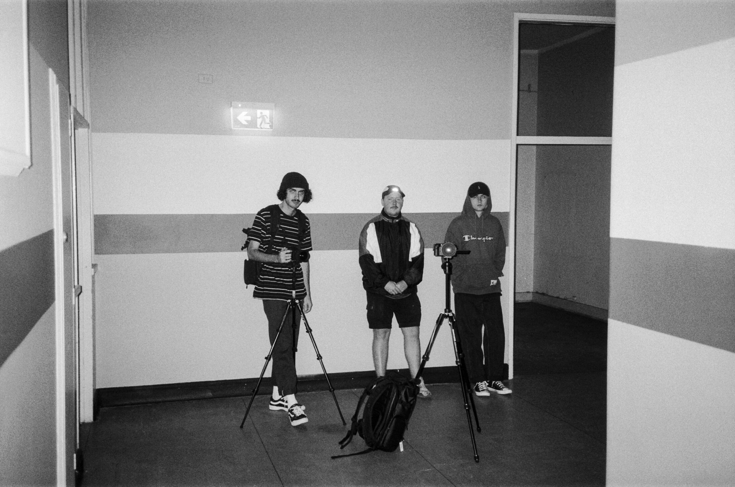 Three young men standing indoors with cameras on tripods, wearing casual streetwear, in front of a wall with an illuminated exit sign.