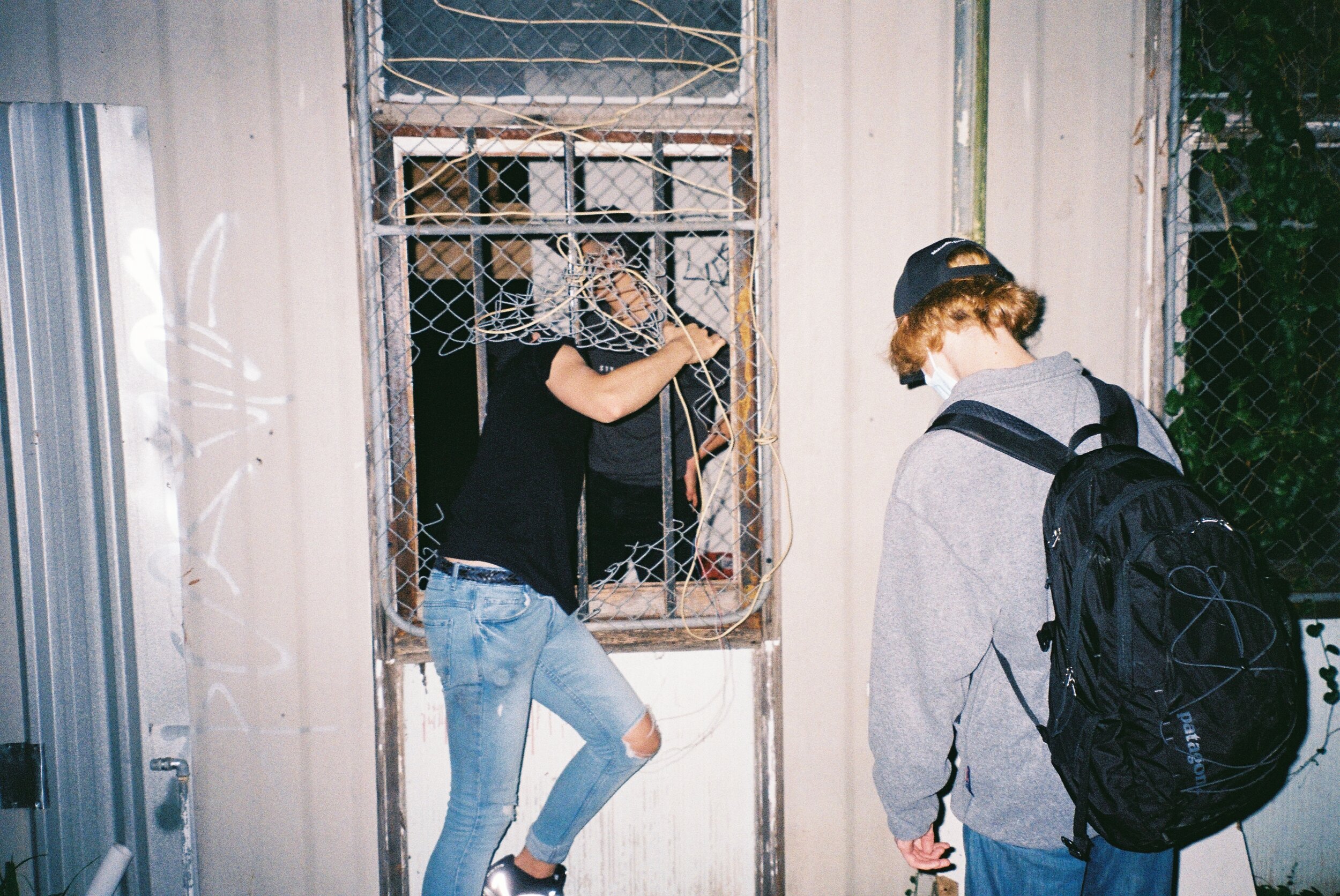 Three teenagers near a damaged wire gate, one girl climbing through, two boys standing outside, one with a backpack, all wearing casual clothing and masks.