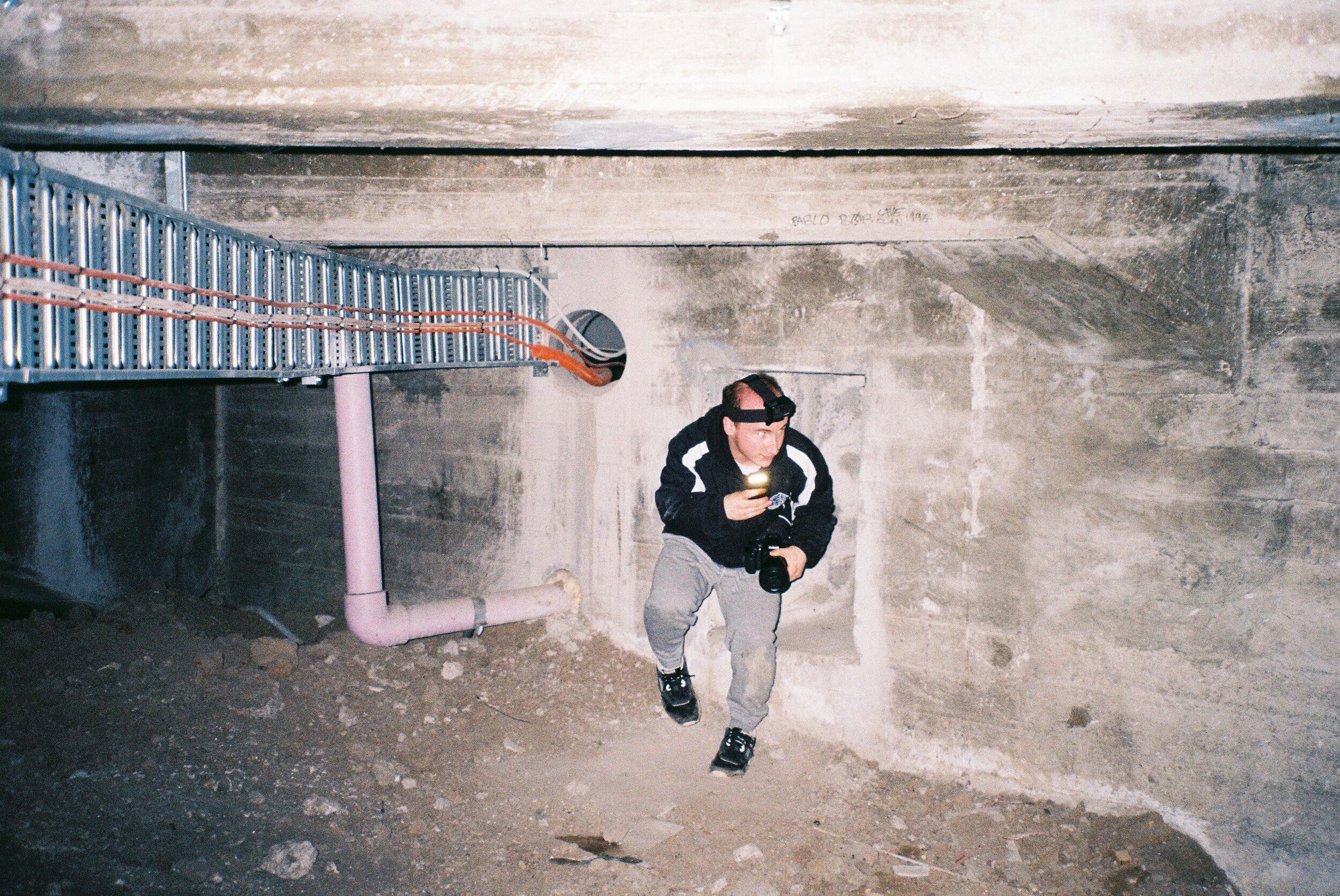 A man with a camera and headlamp, dressed in casual sportswear, is running in a concrete or underground area with exposed pipes and rough walls.