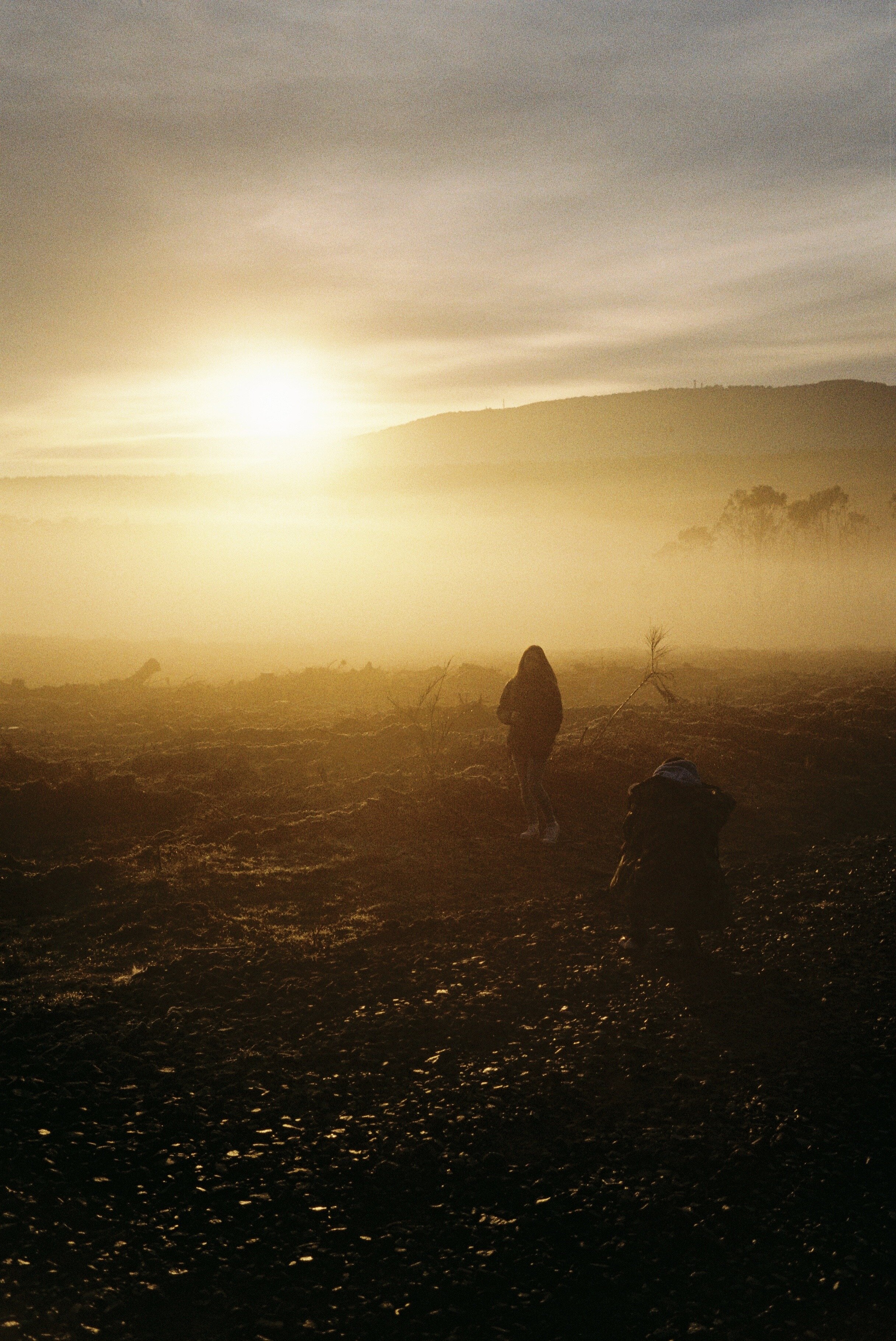Two people in a field during sunrise with hills and mist in the background.