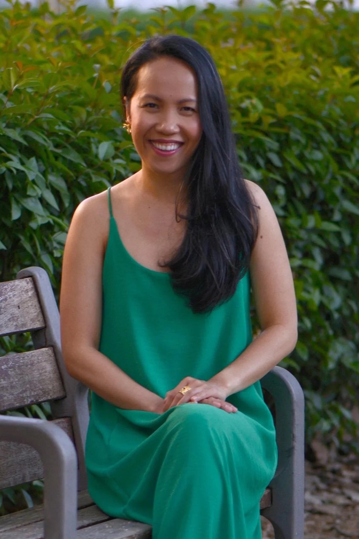 A woman with long black hair sitting on a wooden bench outdoors, smiling, wearing a green dress and gold earrings, with a background of green foliage.