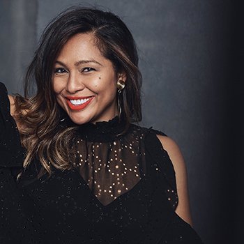 A smiling woman with wavy hair wearing a black dress with sheer, dotted fabric on the shoulders and upper chest.