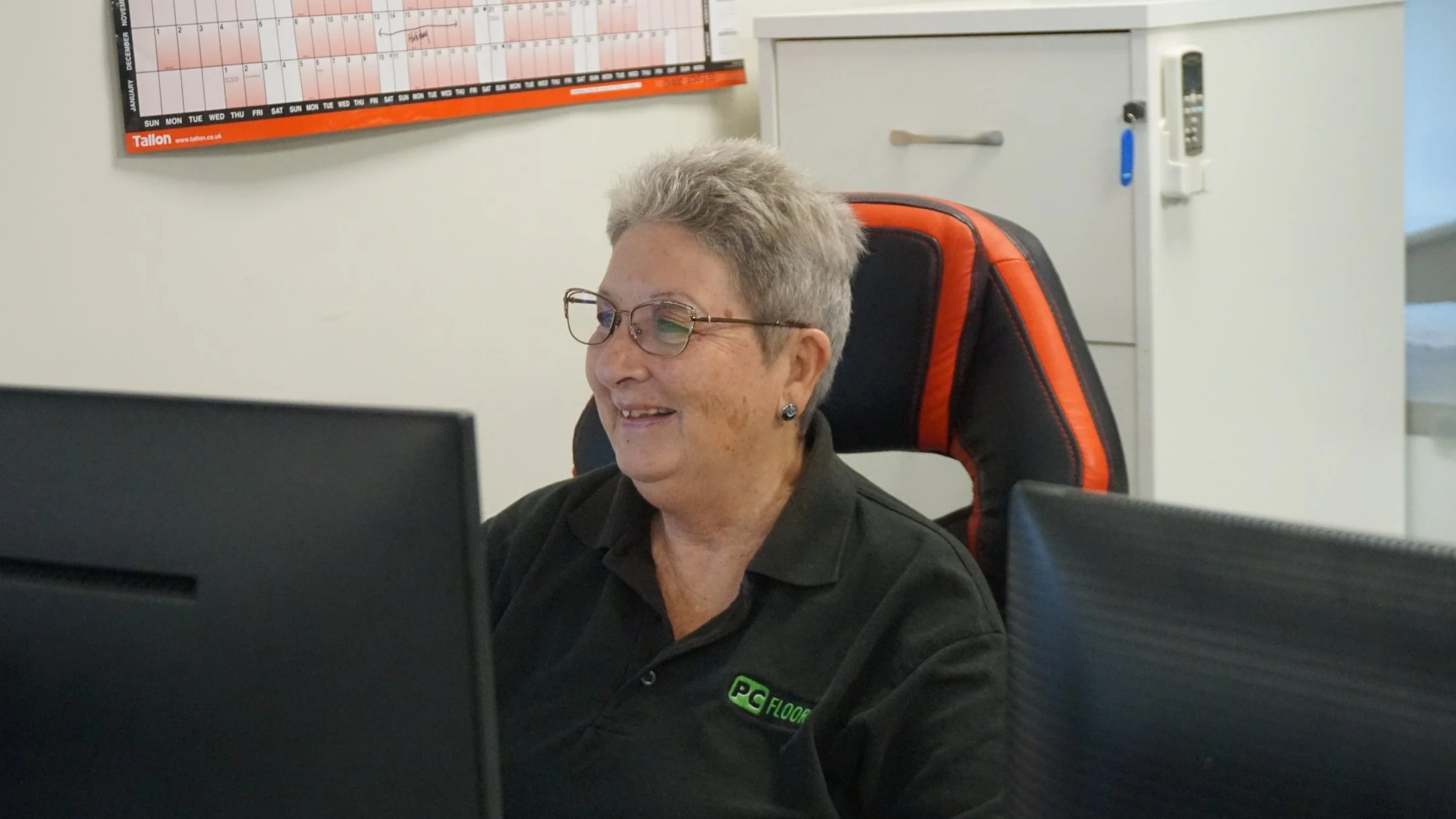 Smiling elderly woman working at a computer in an office, wearing glasses, earrings, and a black polo shirt with a green logo.
