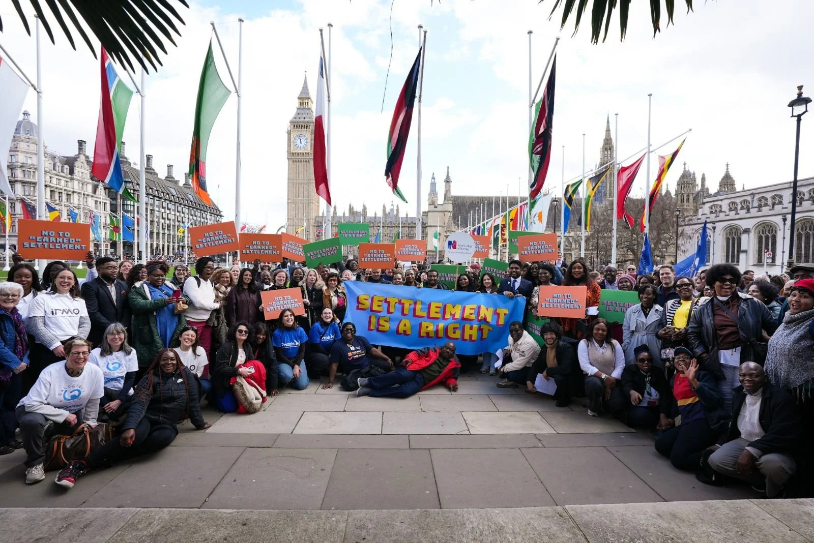 A large group photo of people outside Parliament, holding up colourful signs saying: Settlement is a Right