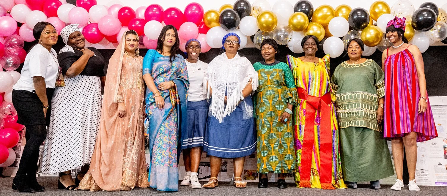 Ten colourfully-dressed women from WAST stand smiling on a stage full of balloons, celebrating WAST's 20th Anniversary.