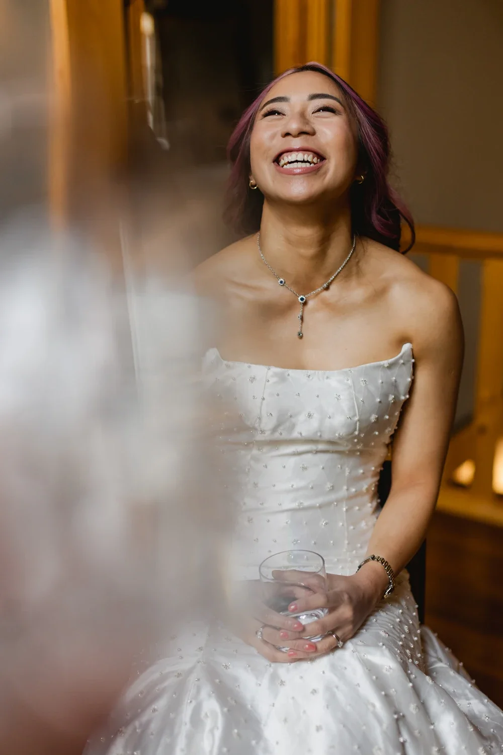 A woman with pink-tinted hair wearing a white dress and jewelry, smiling and holding a glass of water indoors.