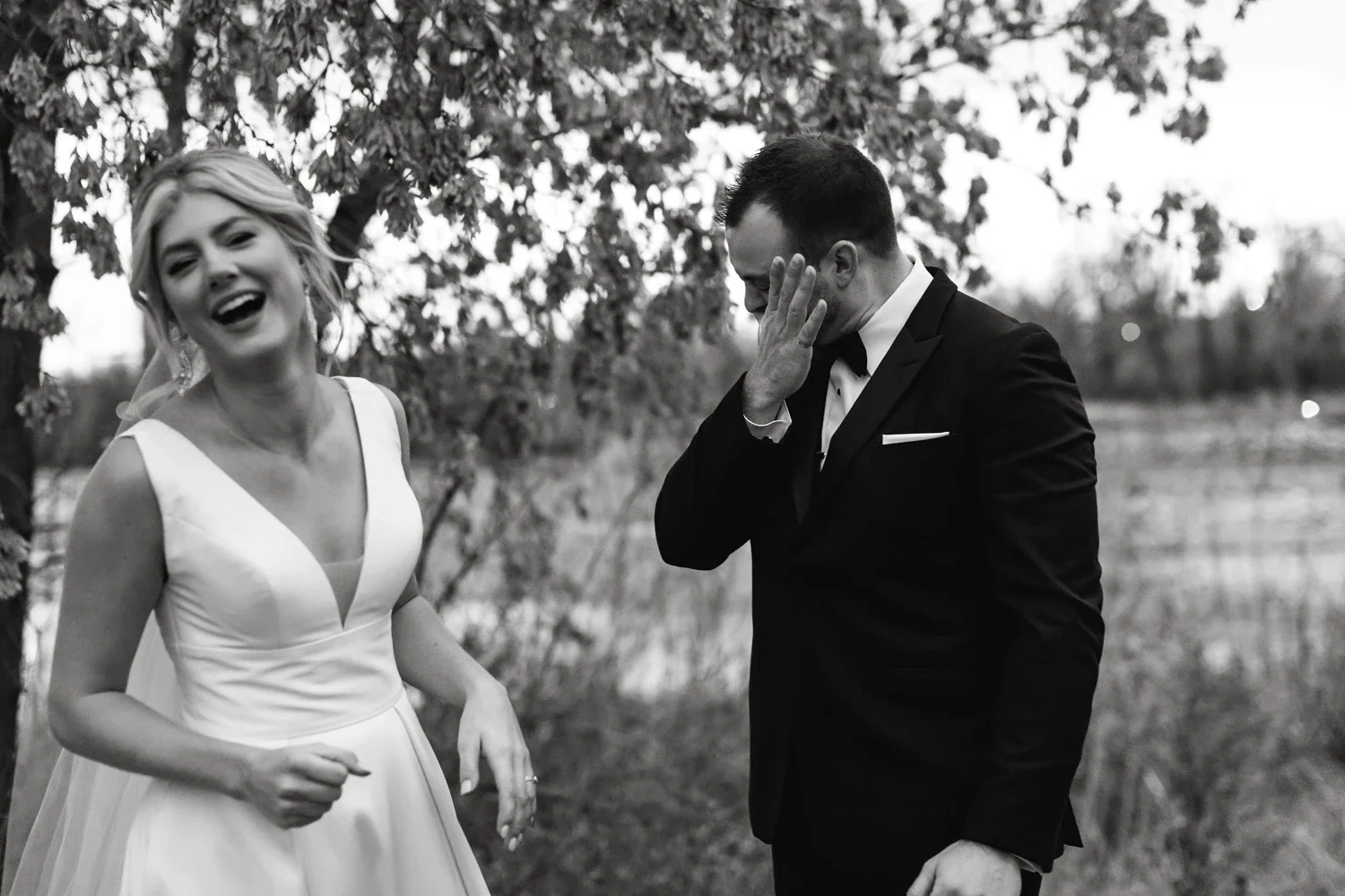 Black and white photo of a bride and groom outdoors, the bride is smiling and laughing, the groom is covering his face with his hand in a moment of emotion or amusement.