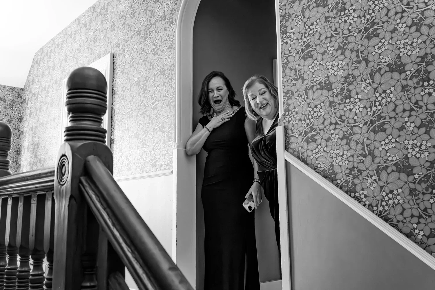 Two women in formal dresses are standing in a doorway, reacting with surprise and amusement.