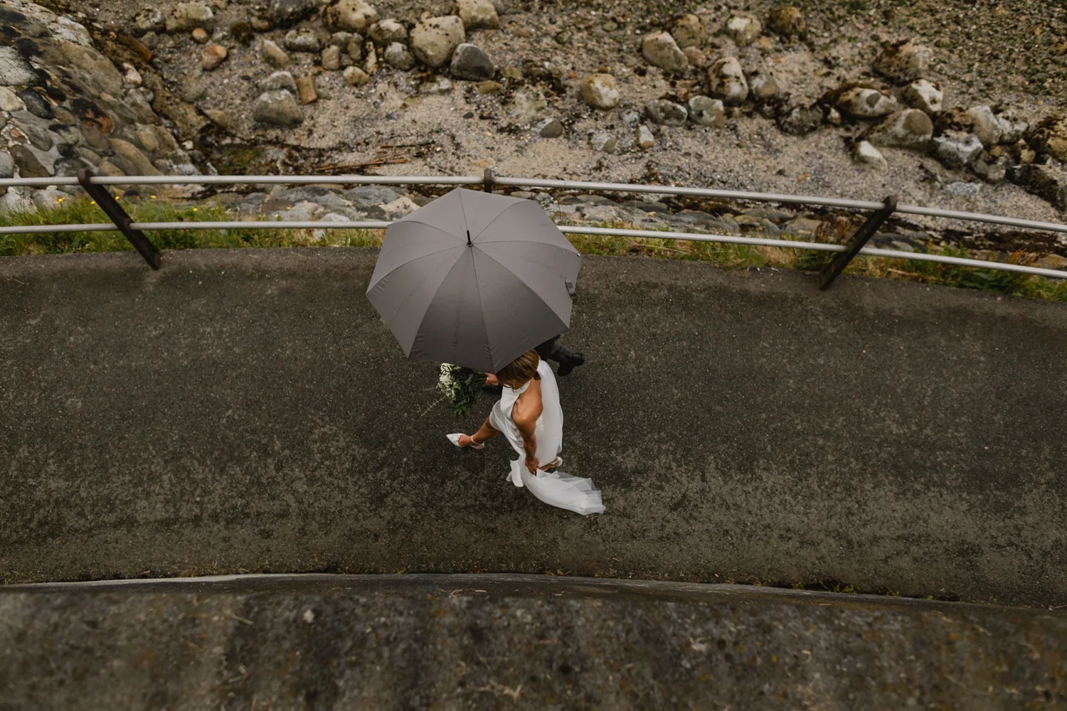 A person holding a white bouquet and a white dress, walking under a gray umbrella on a paved path beside a metal railing, with a rocky surface below.