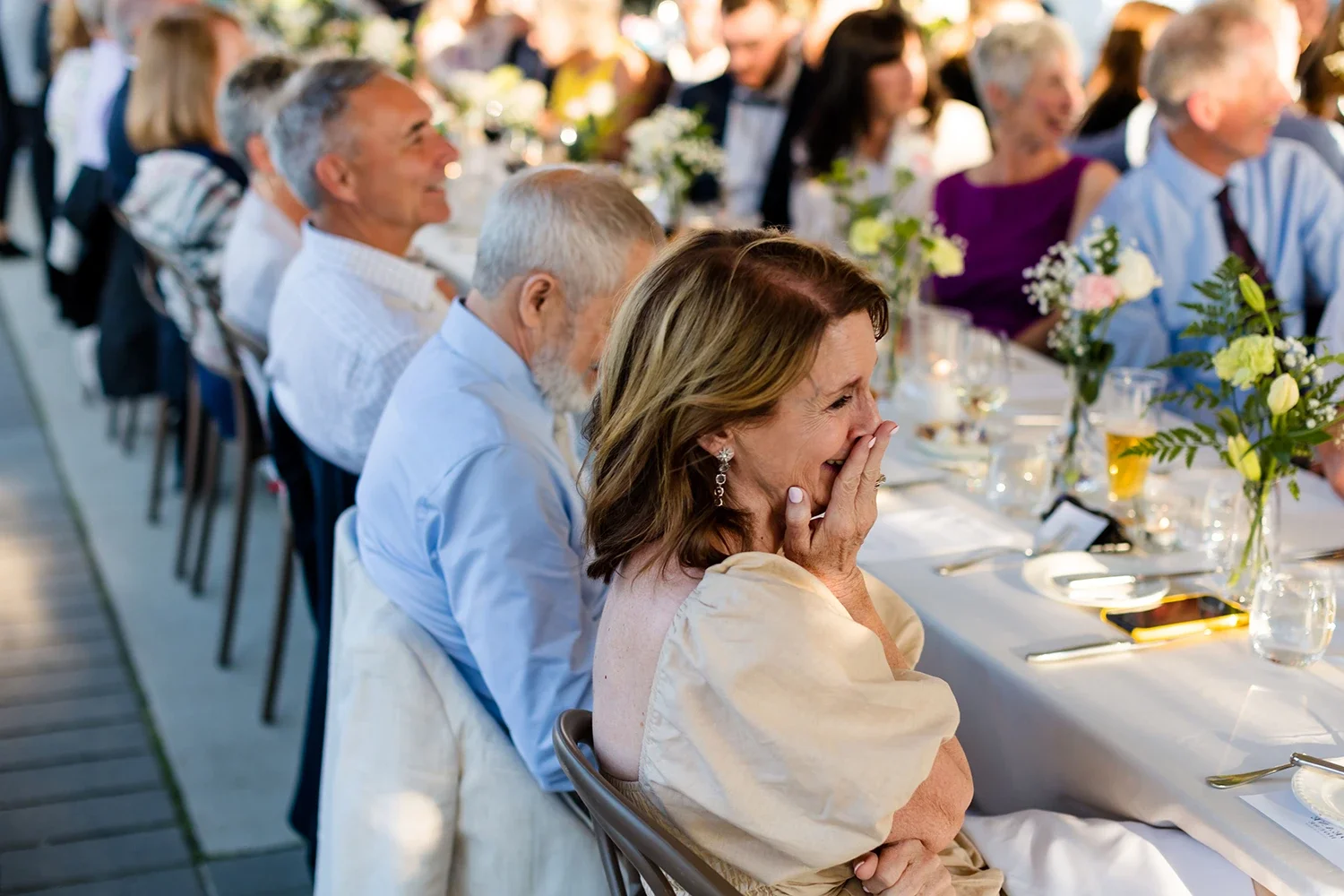 A group of people at a formal event, sitting at a long decorated banquet table, with a woman in the foreground visibly emotional, covering her mouth with her hand and smiling.