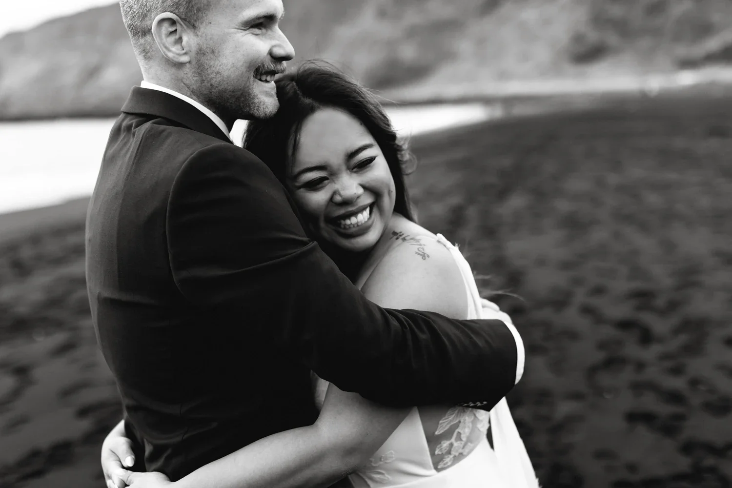 A smiling couple embraces outdoors near a body of water and hills, in black and white.