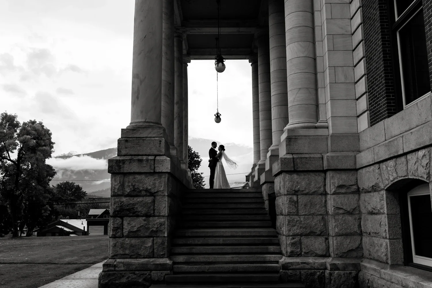 Black and white photo of a bride and groom standing on steps between large marble columns of a building, holding hands and gazing at each other, with trees and mountains in the background.