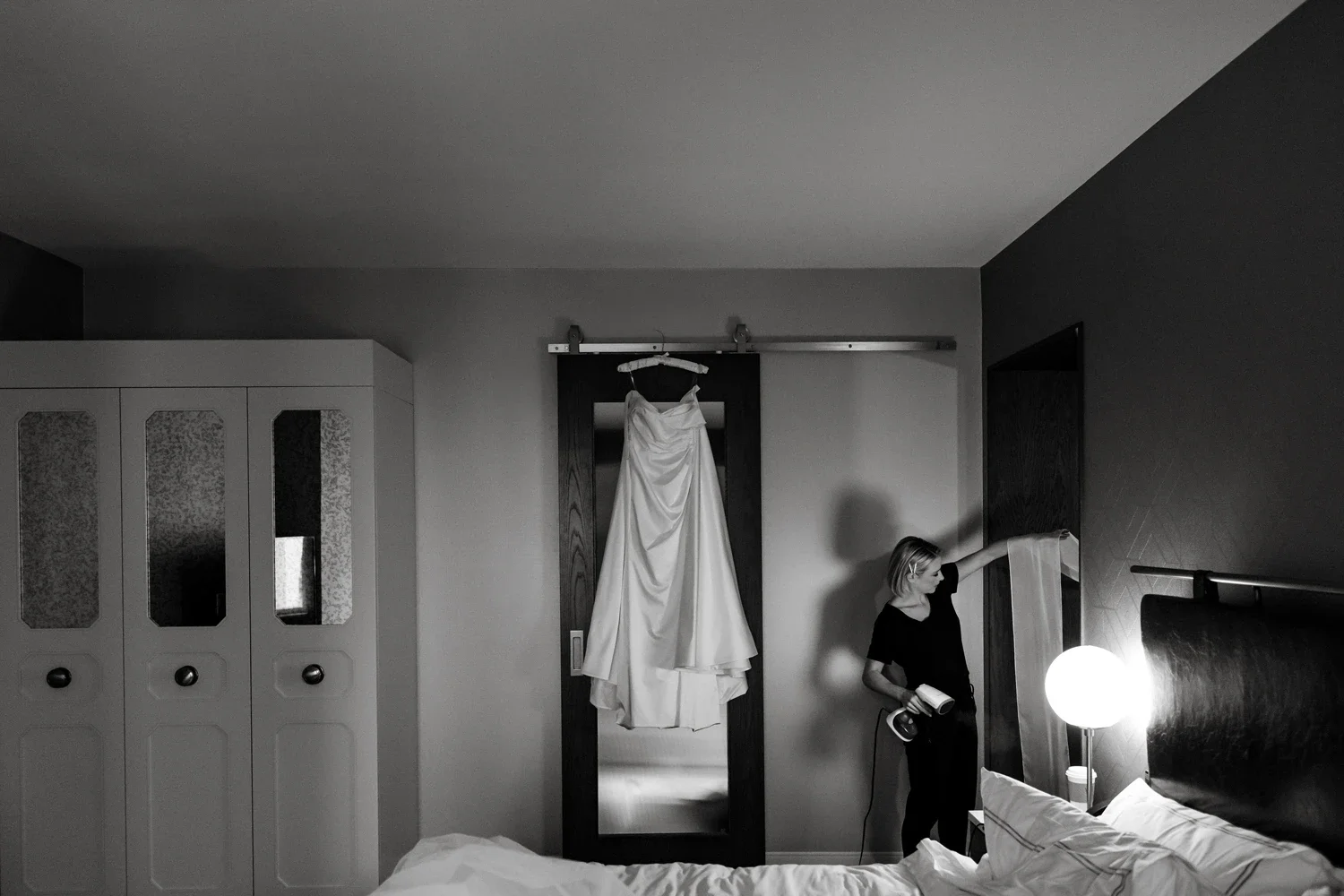 A woman in a black shirt stands next to a bed, holding a hairdryer and brush, in a bedroom with a wedding dress hanging on a mirror door.