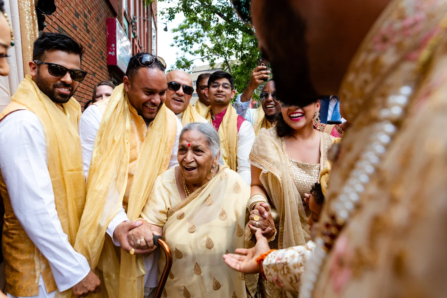 Group of people, dressed in traditional Indian clothing, gathered outdoors smiling and interacting with an elderly woman holding a cane, in a festive celebration.