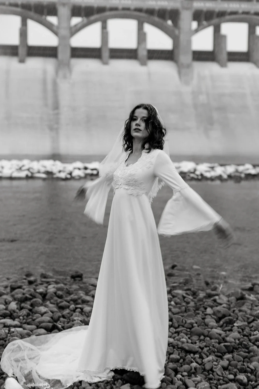 A woman in a long white dress stands on a rocky shore with a bridge in the background.
