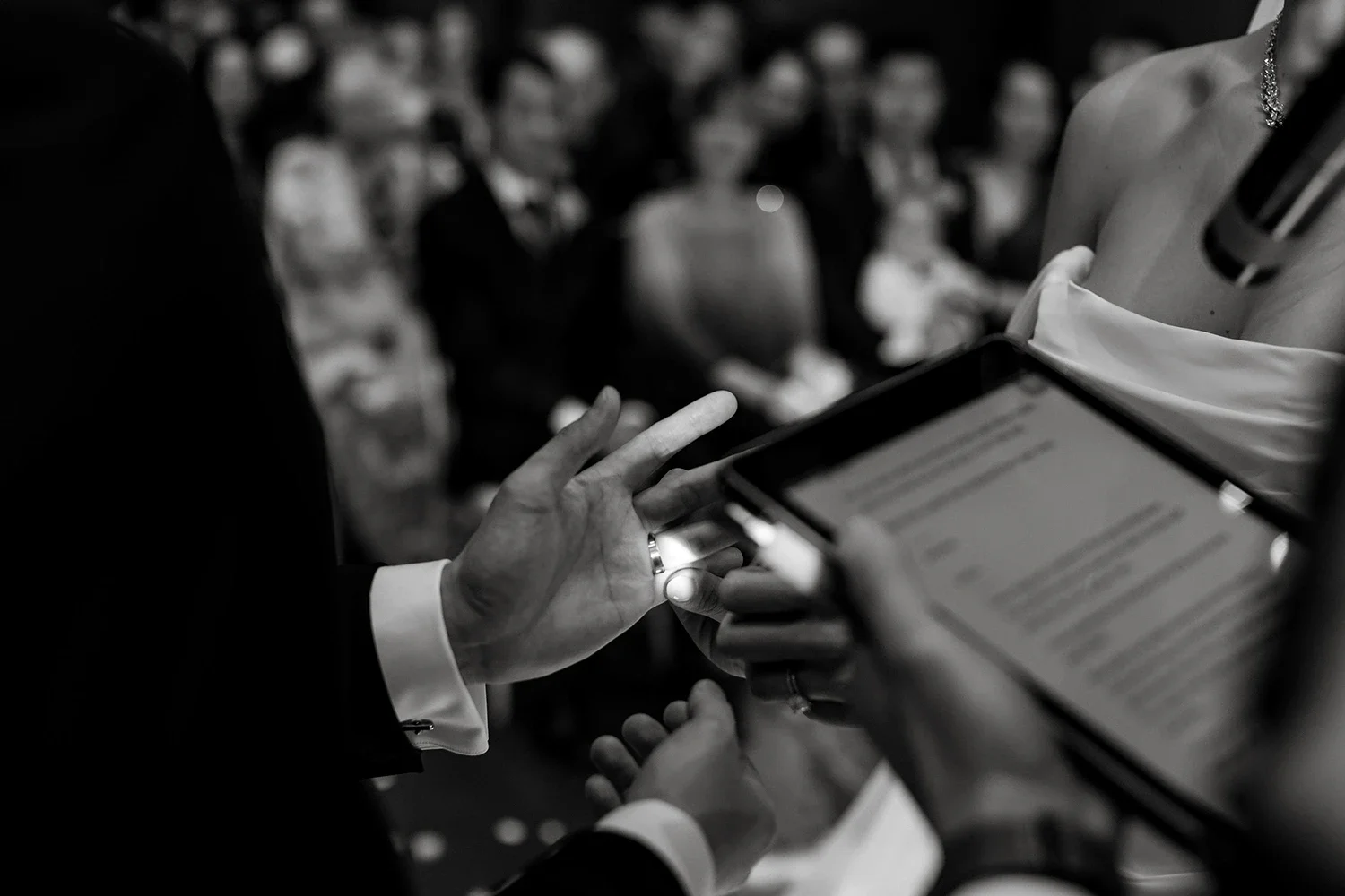 Close-up of a person in formal attire exchanging wedding rings during a wedding ceremony, with an audience in the background.