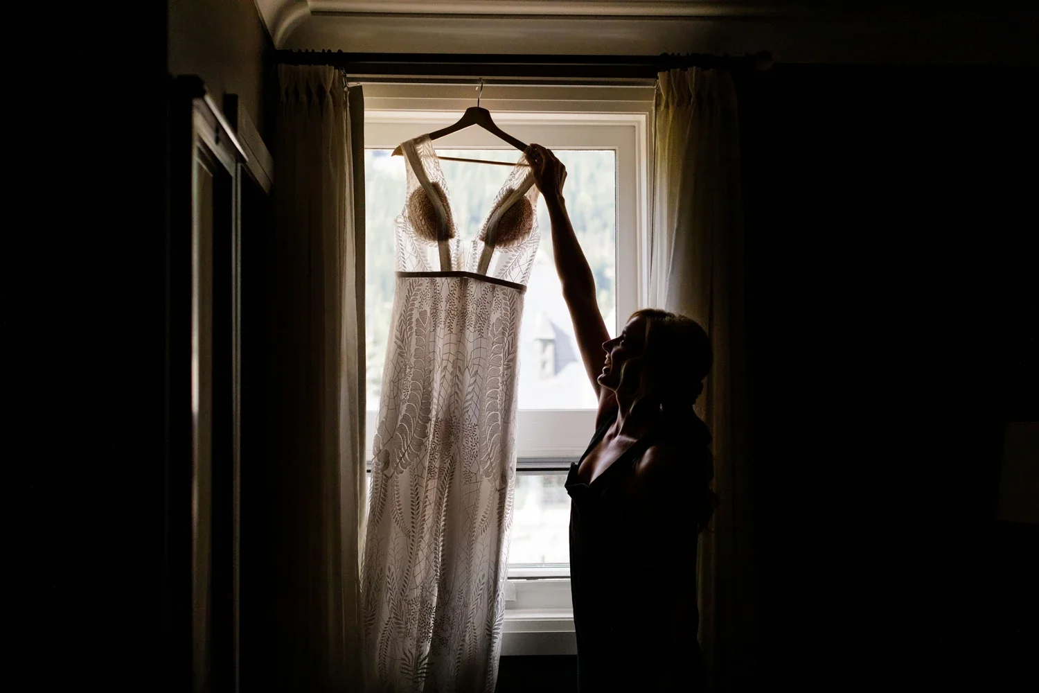 A woman silhouetted in a dark room, holding a wedding dress by a window with light coming through the curtains.