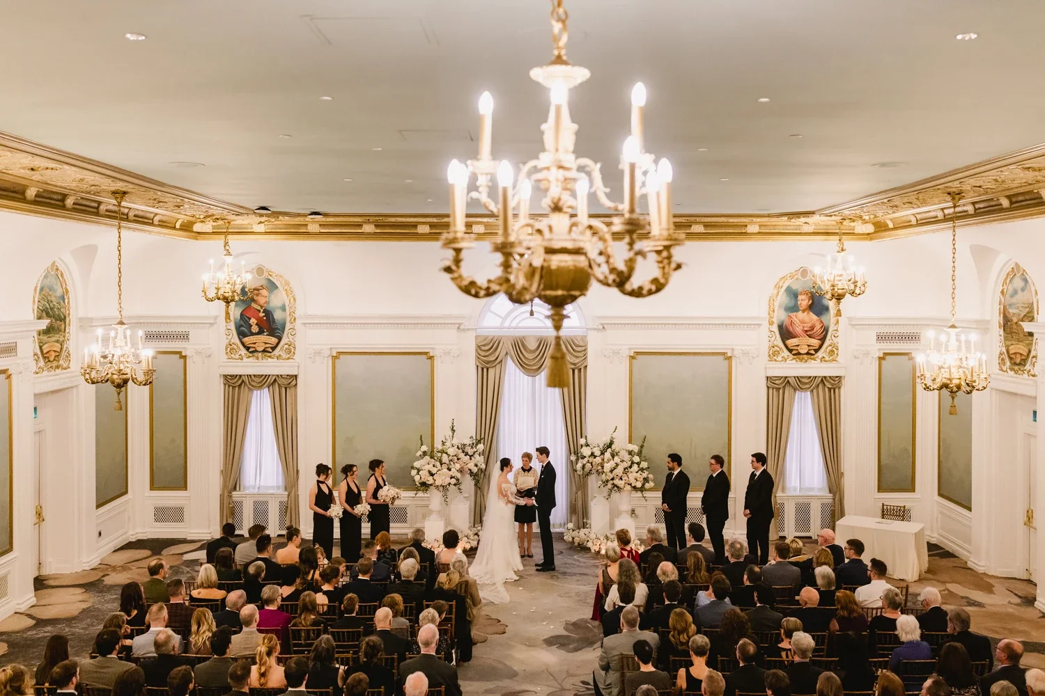 Wedding ceremony in an elegant ballroom with high ceilings, chandeliers, and ornate decorations. The bride and groom are standing in front of an officiant, surrounded by bridesmaids and groomsmen. Guests are seated facing the couple, witnessing the c