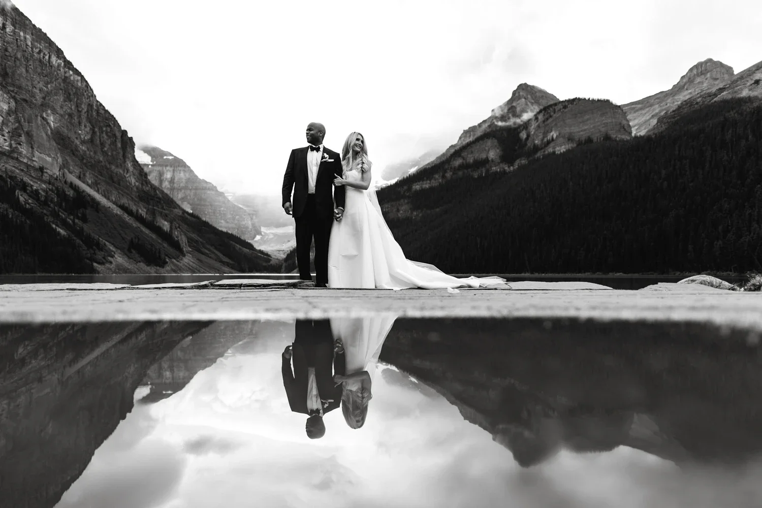 A wedding couple, a man in a tuxedo and a woman in a wedding gown, stands on a dock with mountain scenery in the background. Their reflection is visible in a body of water underneath.