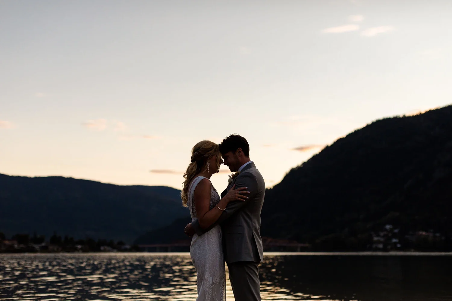 Silhouetted bride and groom embracing by a lake at sunset, mountains in the background.