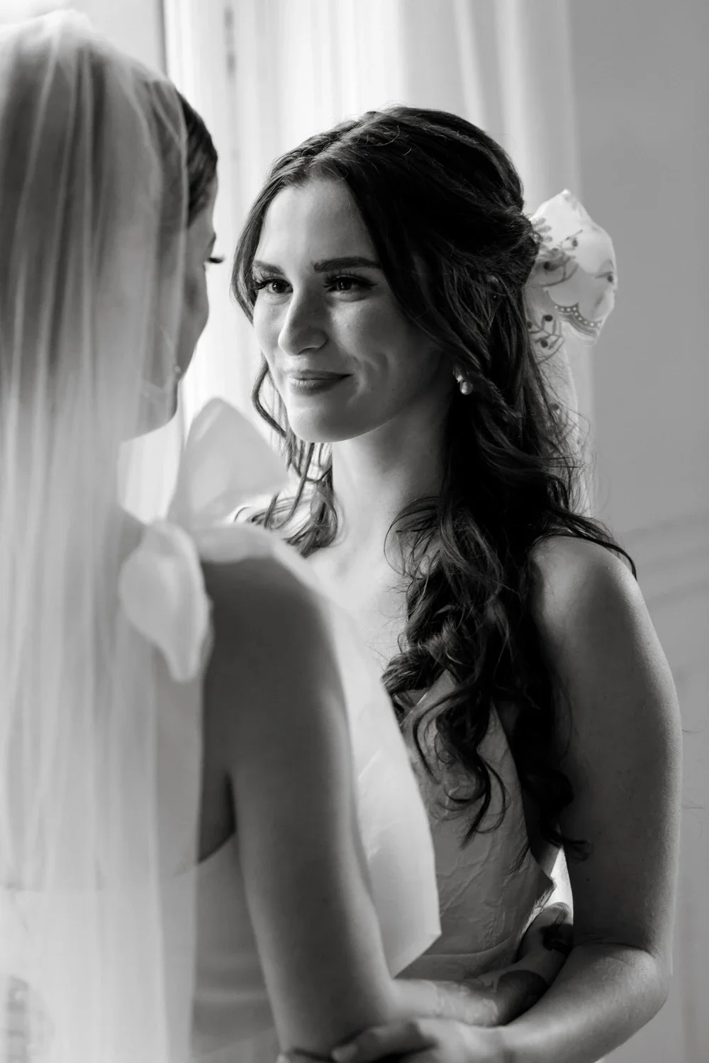Two women, one with long blonde hair wearing a veil, and the other with dark wavy hair and a large floral hair accessory, facing each other closely in a black-and-white photo.