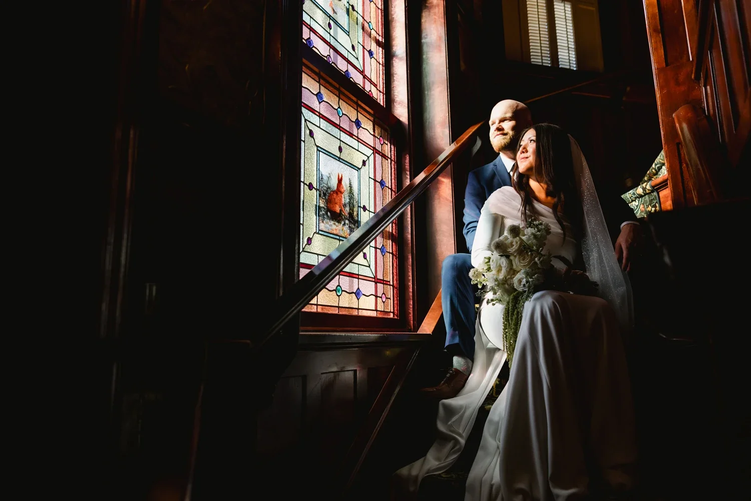 A bride and groom sitting on a staircase near a stained glass window, with the bride holding a bouquet of white flowers and the groom dressed in a navy suit, in a dimly lit, elegant setting.