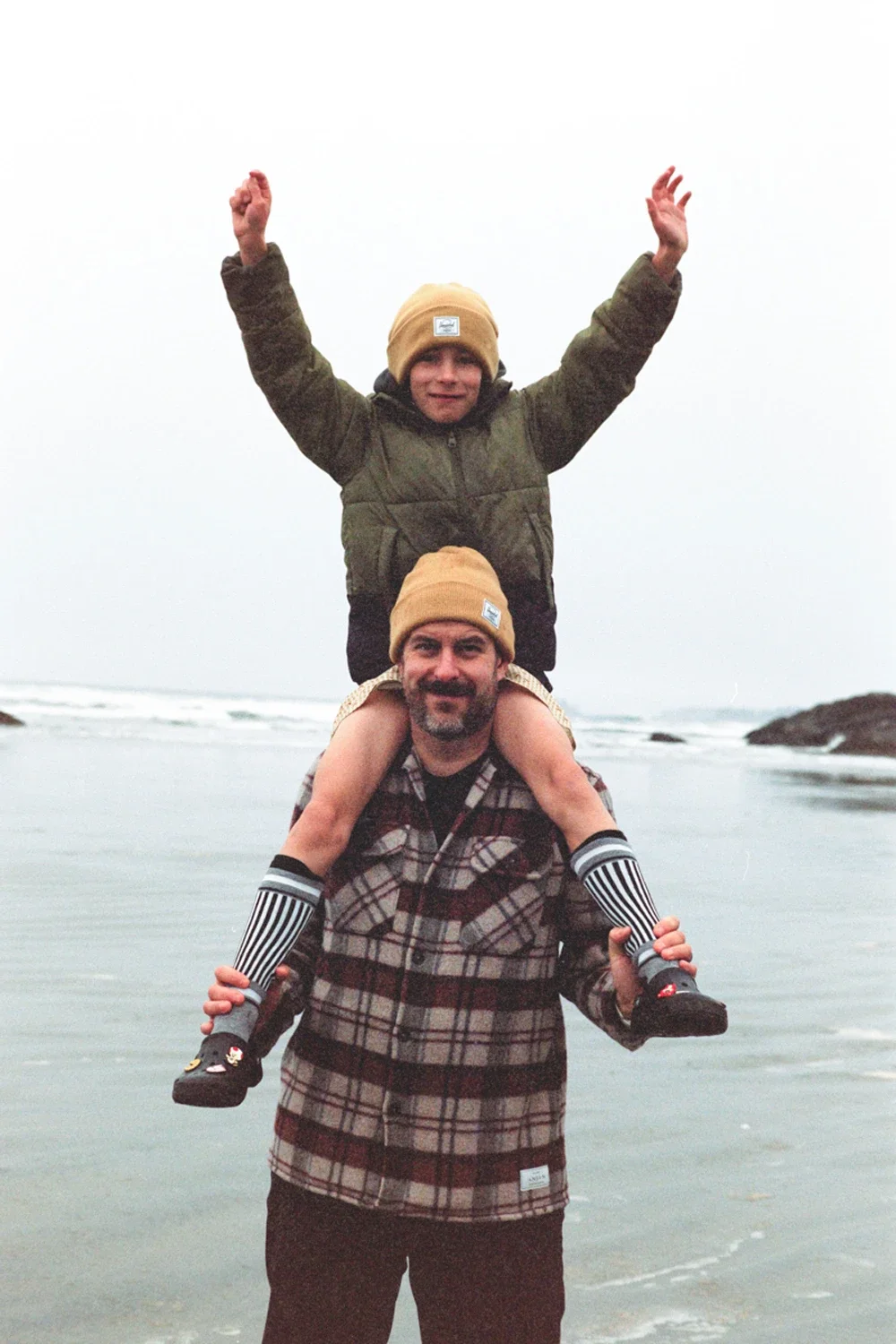 Two men with yellow beanies, one standing and one sitting on his shoulders, at the beach with ocean waves in the background.