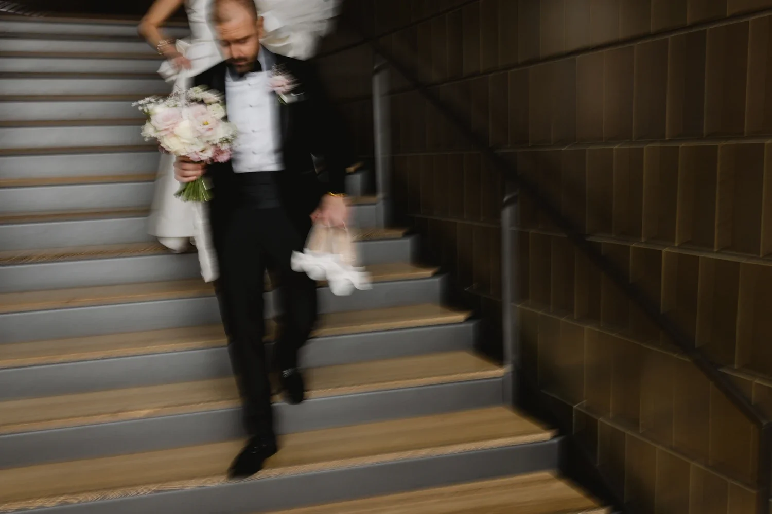 A man wearing a tuxedo with a boutonnière, holding a bouquet of flowers and white shoes, descending a staircase with a blurred background.
