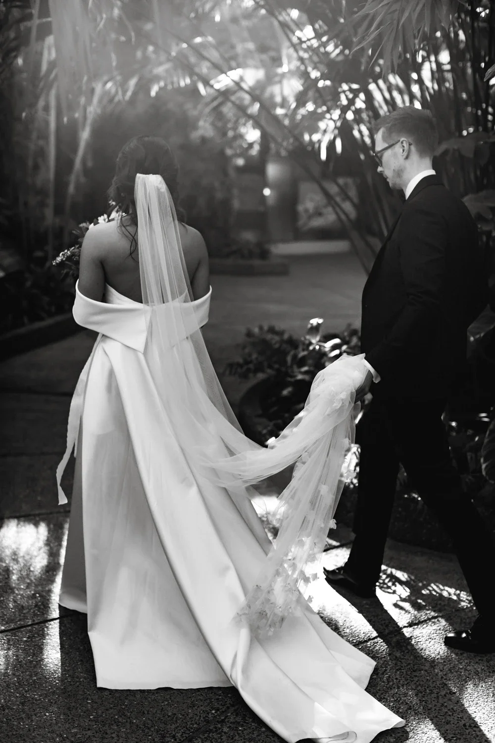 Black and white photo of a bride and groom outdoors, with the groom holding the bride's train. The bride has long hair covered with a veil, and the groom is wearing glasses and a black suit. They are surrounded by large plants, with sunlight filterin