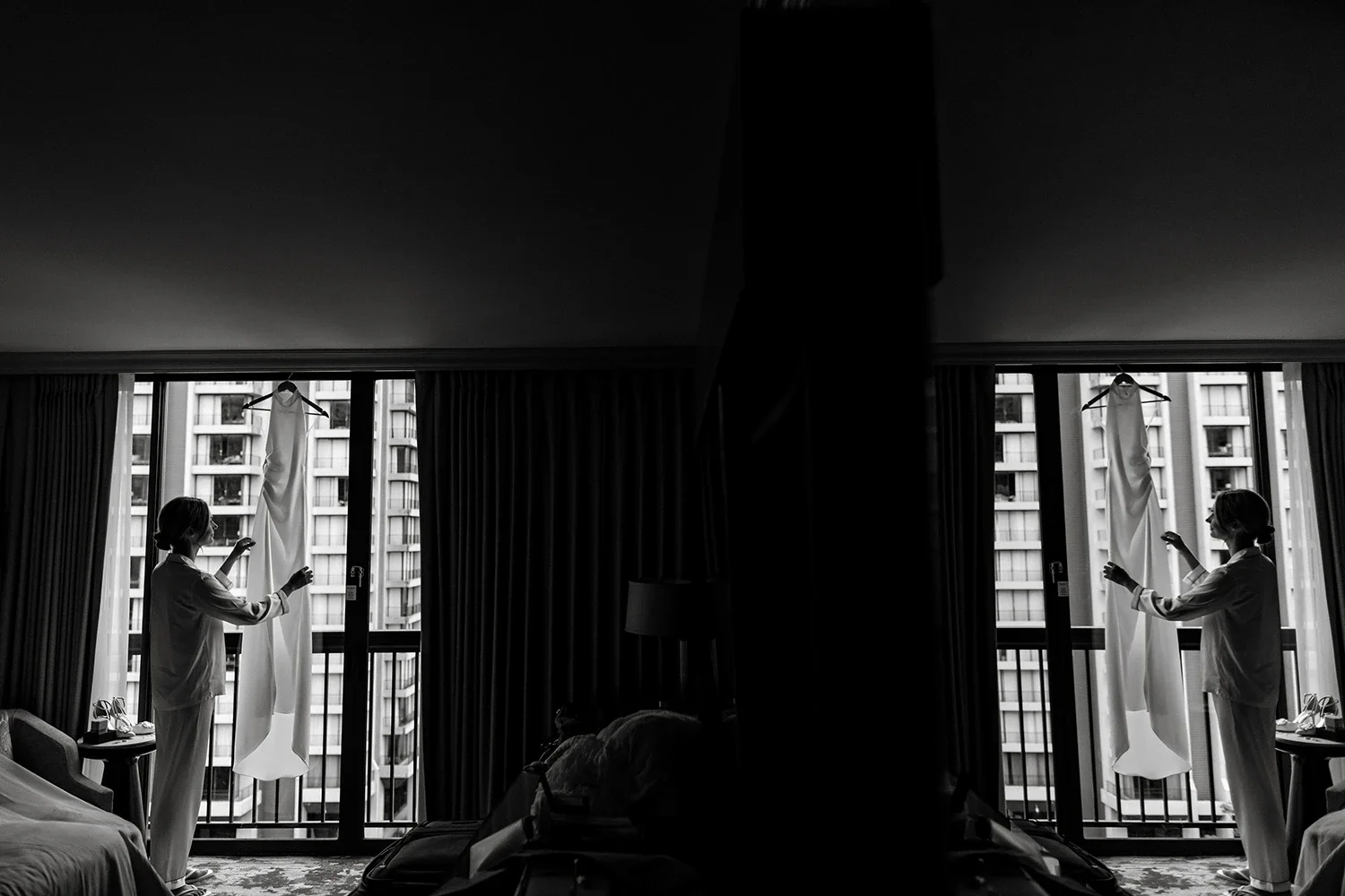 A woman in pajamas standing in front of large windows with curtains, adjusting a wedding dress on a hanger, with a cityscape of tall buildings outside.