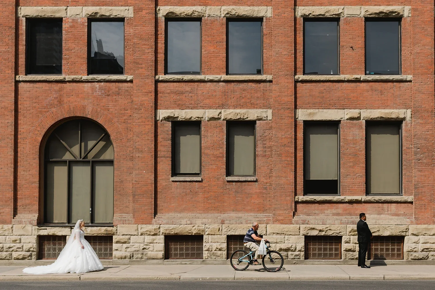 A woman in a white wedding dress walking on a city sidewalk, with a man riding a bicycle, and a man in a black suit standing against a red brick building with six windows.