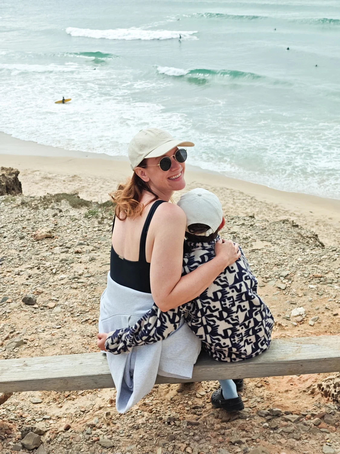 A woman and child sitting on a bench on a beach, embracing, with ocean waves and surfer in the background.