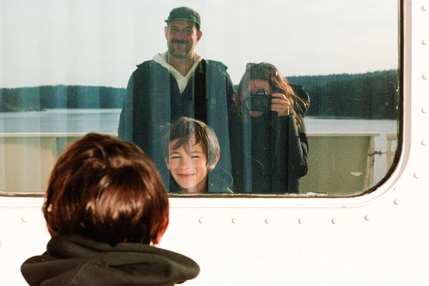 A boy with brown hair takes a photo of himself and two others through a mirror on an airplane window, with a lake and trees in the background.