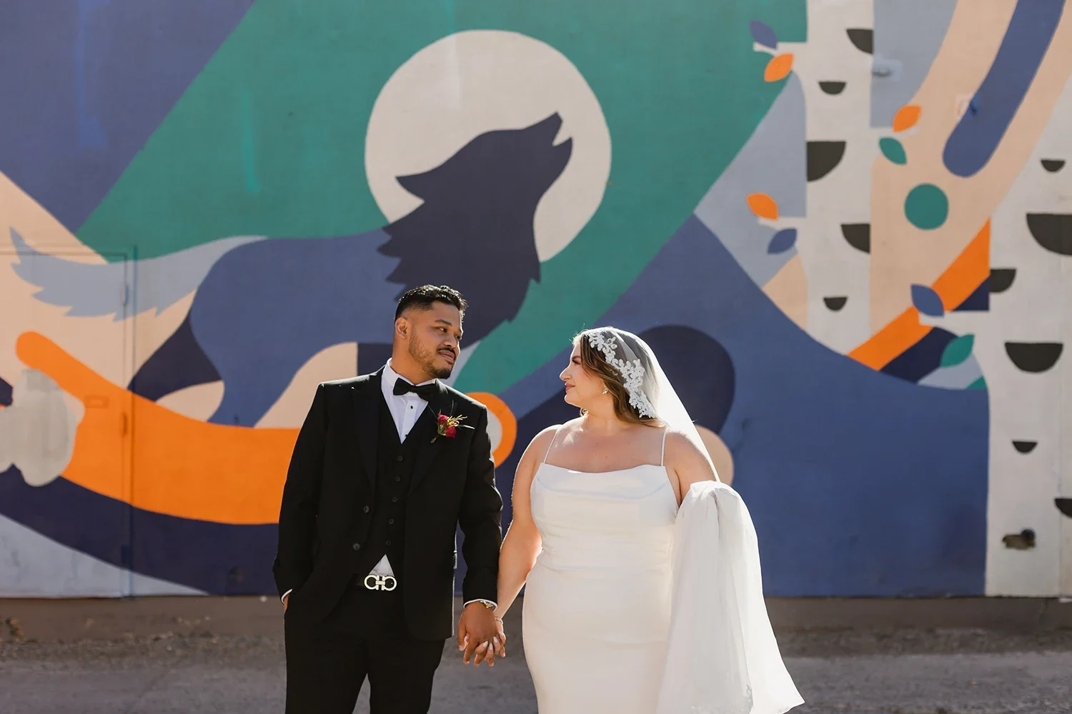 A bride and groom holding hands and looking into each other's eyes in front of a colorful mural featuring a stylized dog and geometric shapes.