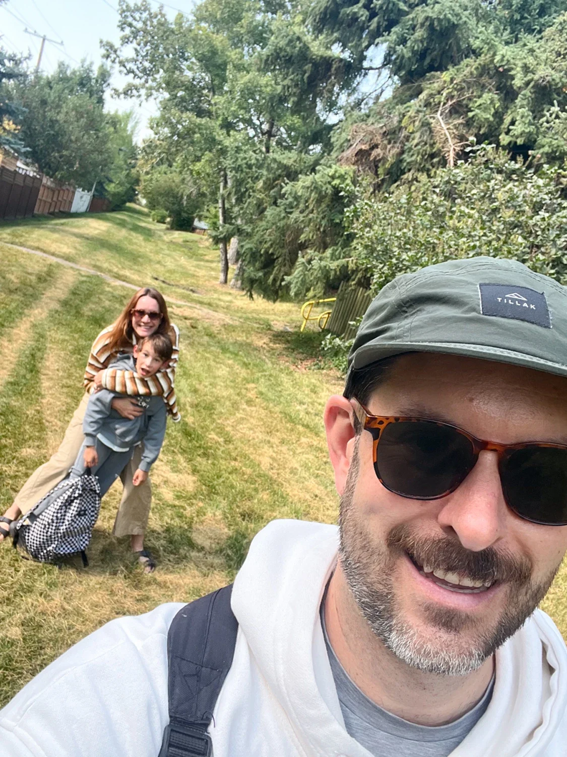 Man taking a selfie with a woman and a boy behind him on a grassy path, trees and fencing in the background.