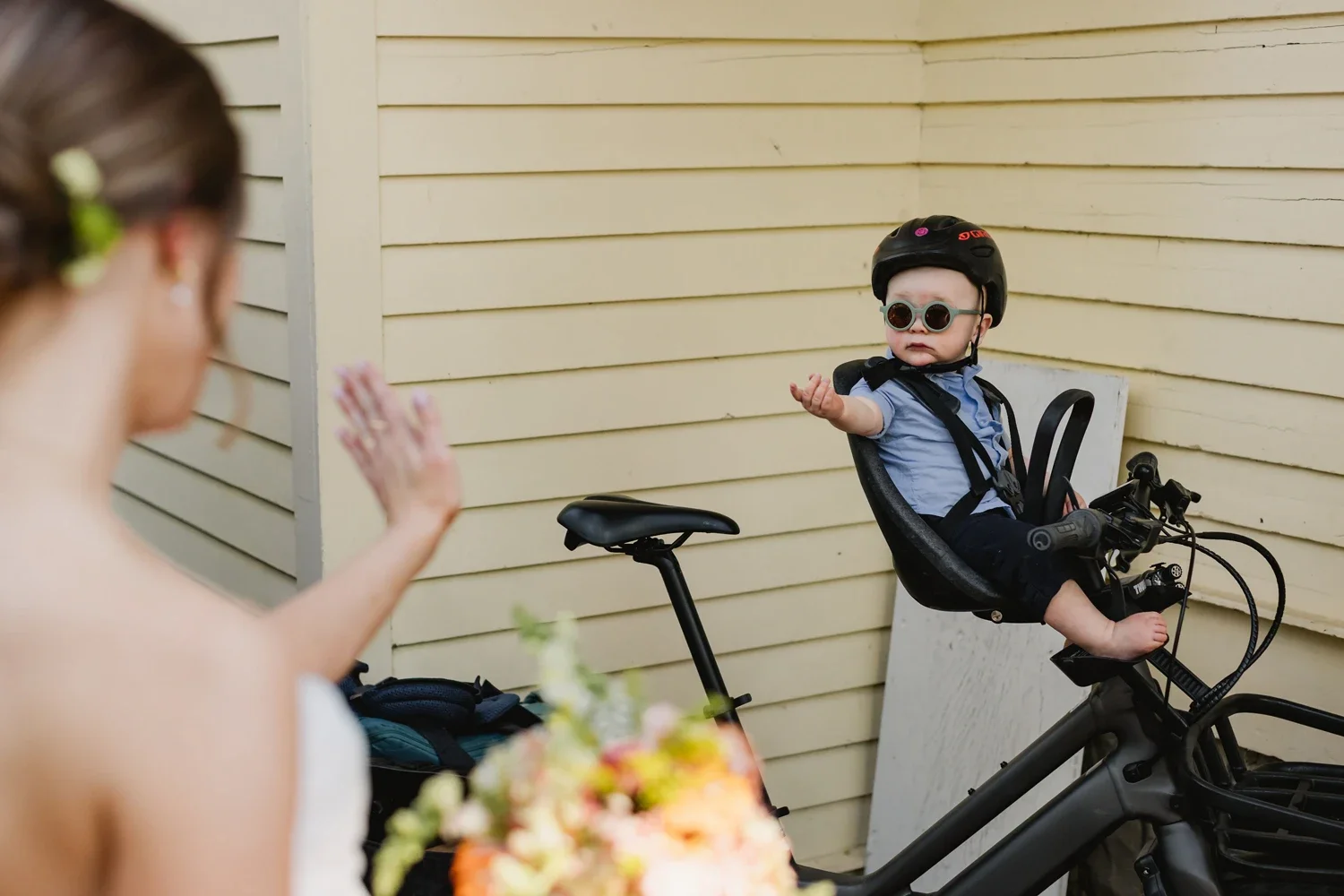 A young child sitting in a child bike seat on a black bicycle, wearing a helmet and sunglasses, reaching out with one hand, in front of a yellow wooden wall.