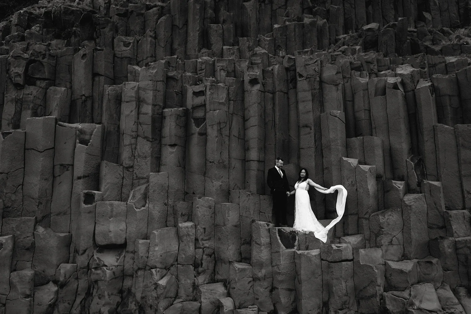 Black and white photo of a couple in wedding attire standing on a rocky cliffside with tall basalt columns in the background.