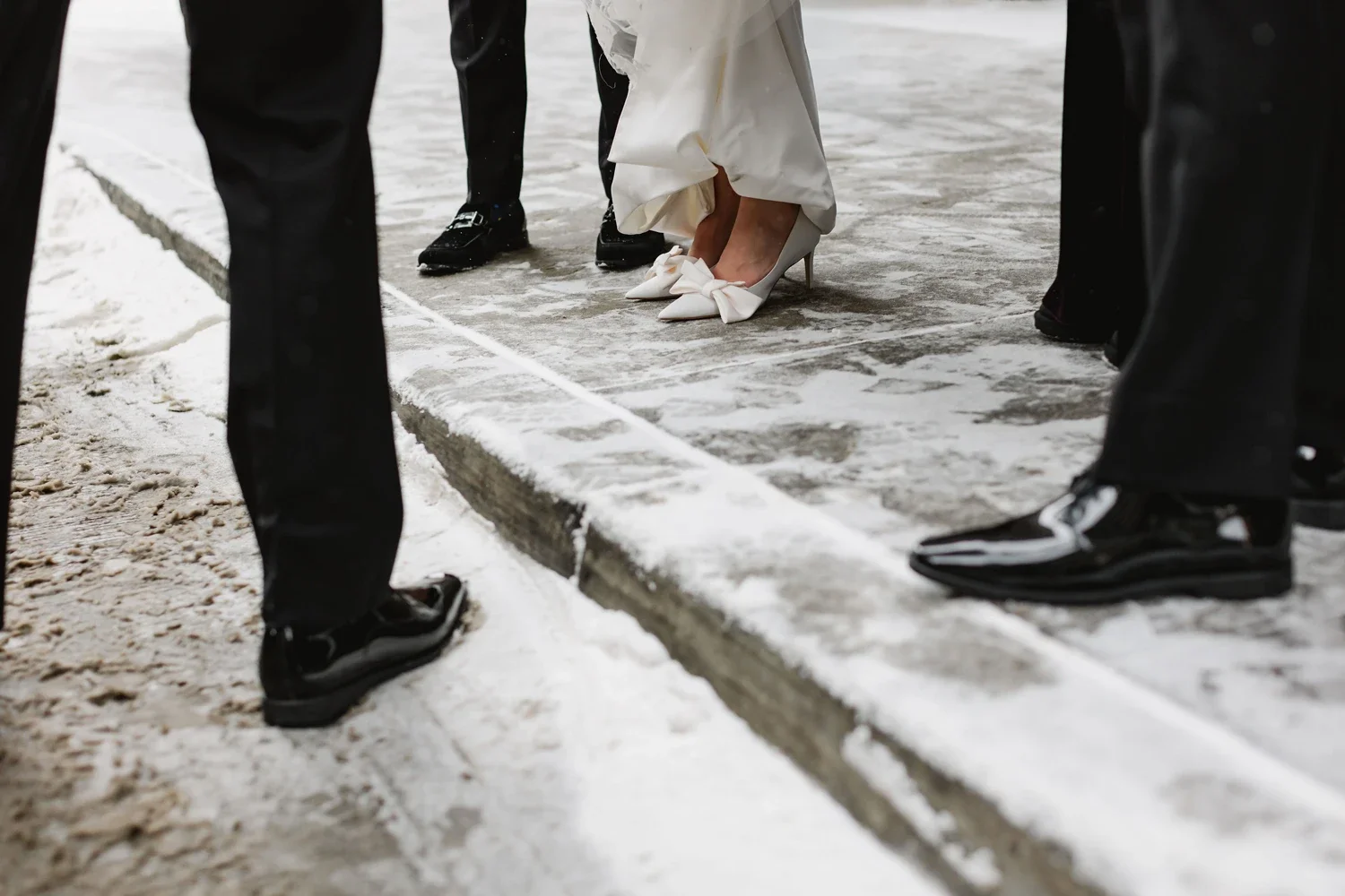 Close-up of a bride wearing white high heels with bows and a wedding dress, standing on a city sidewalk with snowy weather, surrounded by people in formal attire.