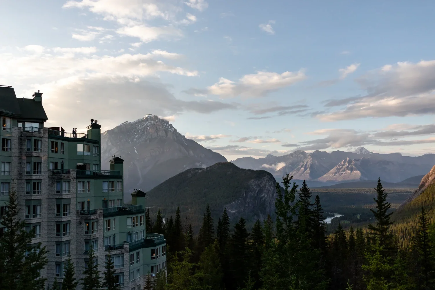 A mountain landscape with snow-capped peaks, a dense forest, and a building in the foreground, during the daytime.