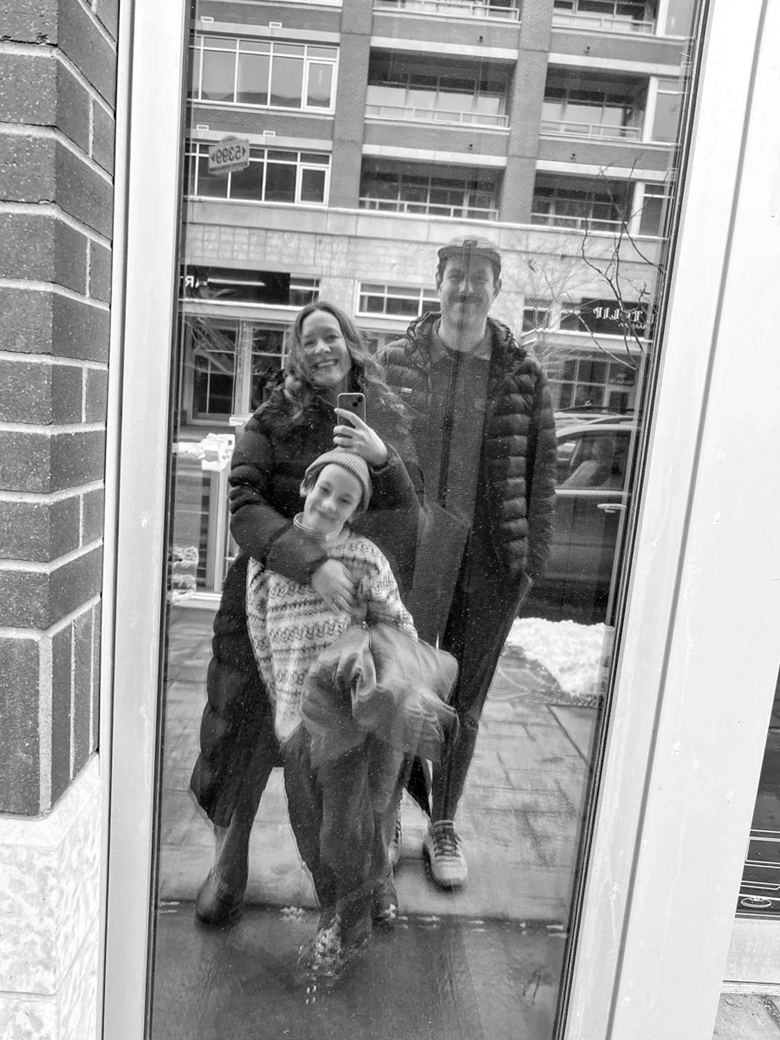 A black-and-white photo of three people taking a mirror selfie outside on a snowy day, with modern apartment buildings in the background. The group includes a woman, a young boy, and a man, all dressed warmly in winter clothing.